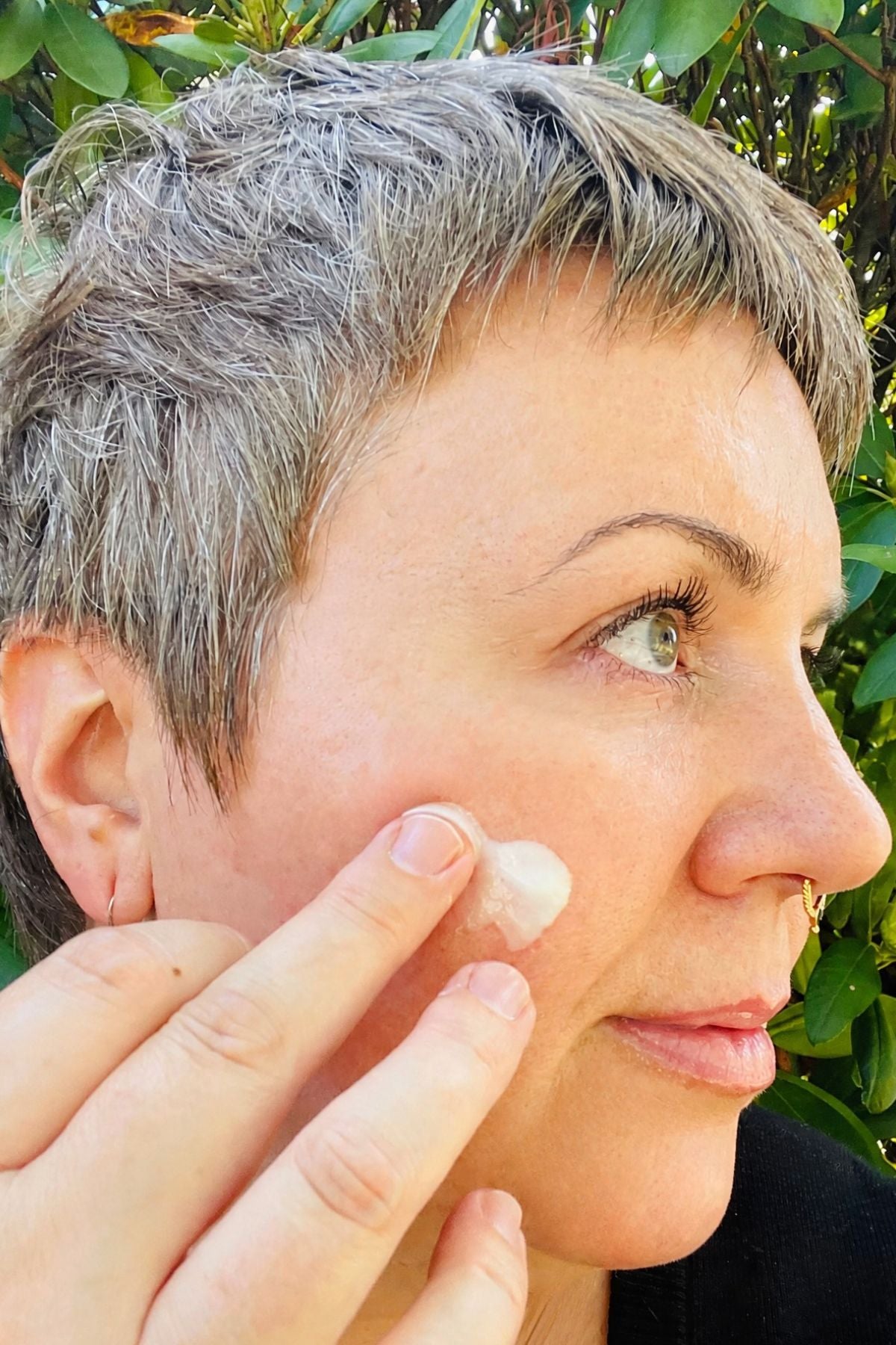 Close-up of a person with short gray hair applying Abracadabra Botanicals A Starry Night's Dream Age-Defying Facial Cream to their cheek, with green foliage in the background.